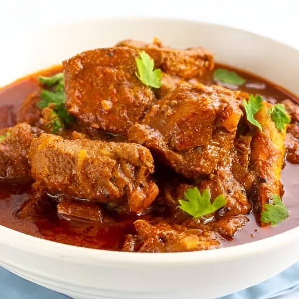 Hearty meat curry in a white bowl, featuring tender pieces and a rich red sauce with green garnish.