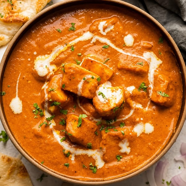 Close-up of a bowl of paneer butter masala with cream swirls, garnished with herbs and naan bread.